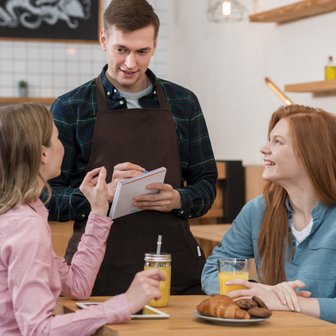 In einem gemütlichen Café diskutieren zwei Freundinnen mit einem Kellner, der ihnen Empfehlungen gibt und Notizen macht.