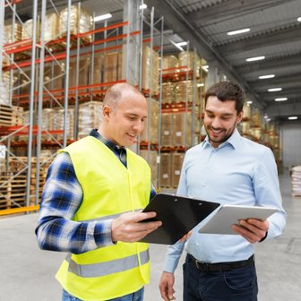 In einem großen Lager diskutieren zwei Männer über ihre Notizen, umgeben von hohen Regalen voller Waren.