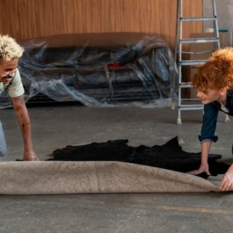 Zwei Personen arbeiten zusammen, um einen großen Teppich in einem modernen Raum auszubreiten, während sie lächeln.