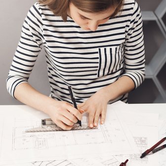 Am Tisch arbeitet eine Frau an technischen Zeichnungen, während sie mit einem Stift präzise Linien auf Papier zieht.