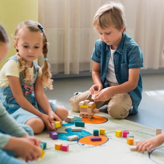 Vier Kinder spielen auf einem bunten Teppich und bauen mit Holzklötzen, während sie konzentriert miteinander interagieren.