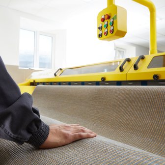 In einem hellen Raum wird ein Stoff von einer Person mit einer dunklen Jacke berührt, während eine Maschine darüber schwebt.