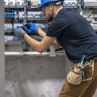 Ein Techniker in einem blauen Helm arbeitet konzentriert an einem Schaltschrank mit vielen Kabeln und Schaltern.