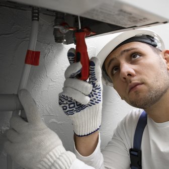 Ein Handwerker mit einem weißen Helm und Handschuhen arbeitet konzentriert an einer Rohrleitung unter einem Waschbecken.