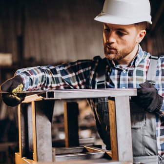 Ein Handwerker in einem grauen Overall und einem karierten Hemd misst präzise ein Metallgestell in einer Werkstatt.