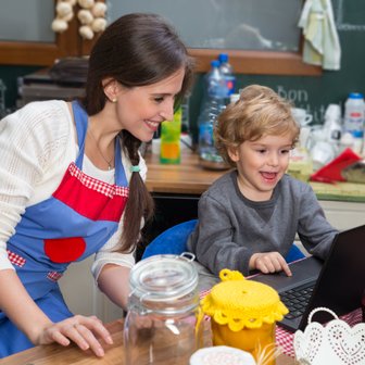 Eine fröhliche Mutter und ihr kleiner Sohn sitzen in der Küche und schauen gemeinsam auf einen Laptop.