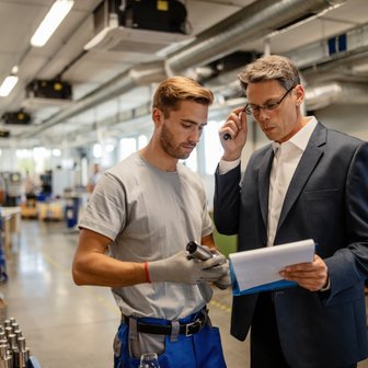 Zwei Männer in einer Produktionsumgebung, einer im Anzug und der andere in Arbeitskleidung, führen ein wichtiges Gespräch.