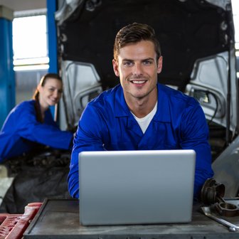 Ein lächelnder Mechaniker sitzt an einem Tisch mit einem Laptop, während eine Kollegin im Hintergrund an einem Auto arbeitet.