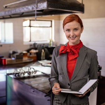 Eine freundliche Köchin mit rotem Halstuch steht in einer gut ausgestatteten Küche und notiert wichtige Informationen.