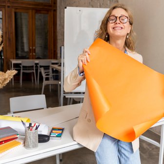 Mit einem breiten Lächeln hält eine Frau ein großes orangefarbenes Papier, während sie in einem hellen, modernen Büro arbeitet.