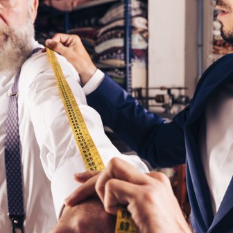 In einem Atelier wird ein älterer Herr von einem Schneider vermessen, um einen maßgeschneiderten Anzug zu erstellen.