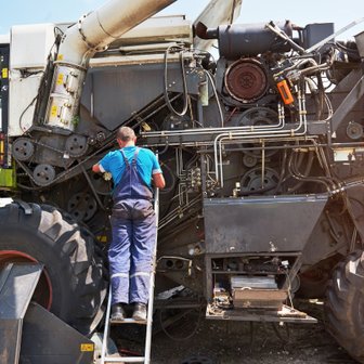 Ein Mechaniker steht auf einer Leiter und repariert die Maschine eines großen Erntefahrzeugs unter blauem Himmel.