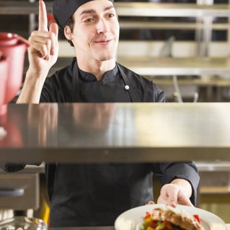 Der Koch hebt einen Finger, während er einen Teller mit einer köstlichen Mahlzeit in einer professionellen Küche reicht.