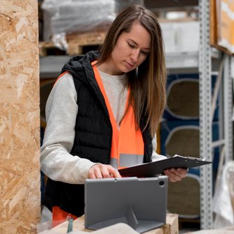 In einem gut organisierten Lager überprüft eine Frau mit einem Tablet und einem Notizblock ihre Arbeitsaufgaben.