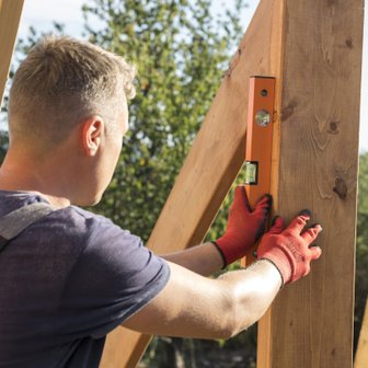 Ein Handwerker überprüft mit einer Wasserwaage die Ausrichtung eines Holzpfostens in einem sonnigen Garten.