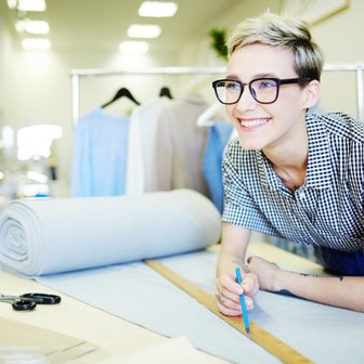In einem modernen Nähatelier plant eine junge Frau mit einem Bleistift auf einem Stoff, umgeben von verschiedenen Kleidungsstücken.