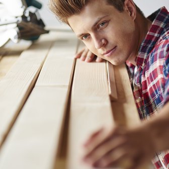 Ein Handwerker in einem karierten Hemd untersucht aufmerksam die Holzlatten, die vor ihm auf einem Tisch liegen.