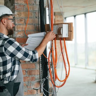 Ein Bauarbeiter mit Helm und Schutzbrille überprüft Pläne an einer Wand mit freiliegenden Kabeln in einem Rohbau.
