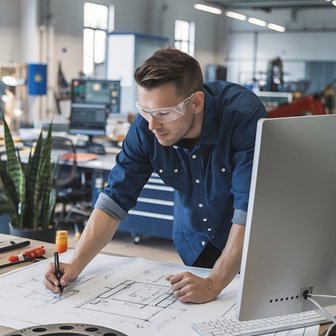 In einer modernen Werkstatt arbeitet ein Mann an technischen Zeichnungen, umgeben von Maschinen und Werkzeugen auf dem Tisch.