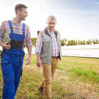 In einem ländlichen Umfeld unterhalten sich zwei Männer, einer trägt blaue Arbeitskleidung und der andere eine Weste.