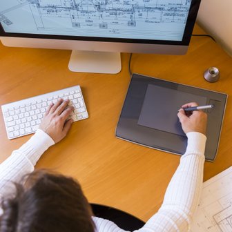 Ein Mensch arbeitet an einem Schreibtisch, nutzt ein Grafiktablett und schaut auf einen Computerbildschirm mit technischen Zeichnungen.