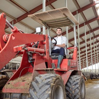 Der Landwirt lächelt, während er auf dem Traktor sitzt, umgeben von landwirtschaftlichen Maschinen und einem sauberen Stall.