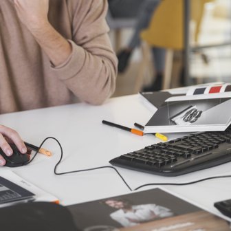 Auf dem Tisch liegen verschiedene Stifte und Zeitschriften, während die Person konzentriert am Computer arbeitet.