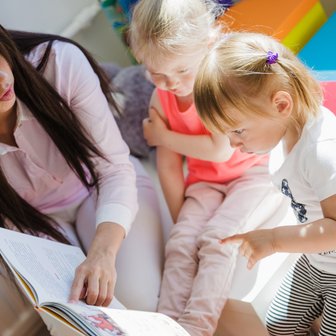 Die Szene zeigt eine Lesestunde, in der eine Frau mit zwei Kindern interagiert, umgeben von bunten Spielmaterialien.