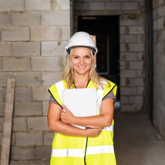 Eine lächelnde Frau in einem gelben Sicherheitsweste und Helm steht in einem Rohbau und hält ein Dokument in den Händen.