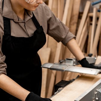 Eine Frau in Schutzbrille und Handschuhen bearbeitet ein Holzstück an einer Maschine in einer Werkstatt voller Holz.