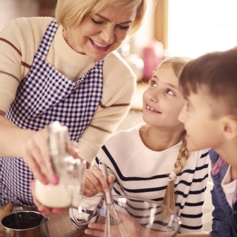 Eine lächelnde Frau in einer karierten Schürze gießt Zucker in eine Schüssel, während zwei Kinder gespannt zuschauen.
