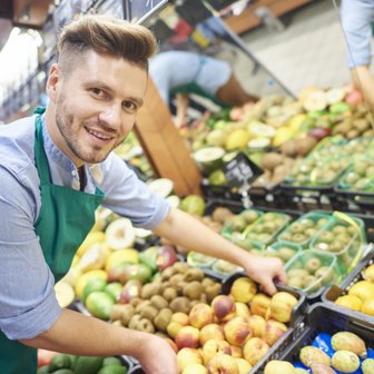 Ein junger Mann mit einem grünen Schürze lächelt, während er frisches Obst in einem gut gefüllten Supermarktregal arrangiert.