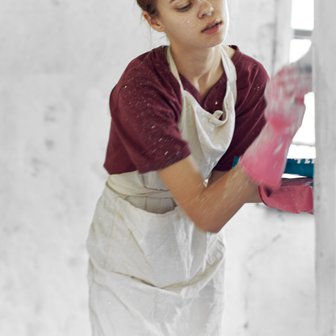 Eine junge Frau in einem schmutzigen Raum putzt mit pinken Gummihandschuhen eine Wand, während sie konzentriert aussieht.