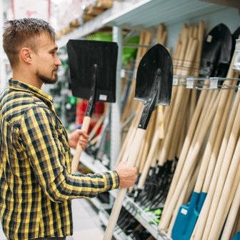 Ein Mann mit einem gestreiften Hemd steht in einem Baumarkt und vergleicht zwei Schaufeln, die er in der Hand hält.