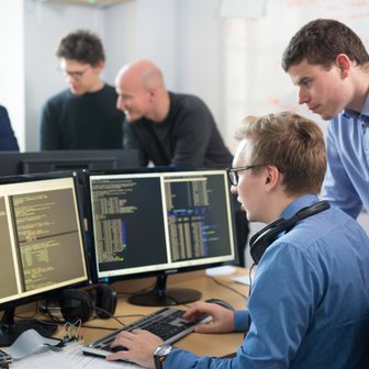 Ein Programmierer arbeitet konzentriert an zwei Bildschirmen, während Kollegen im Hintergrund über den Code diskutieren.