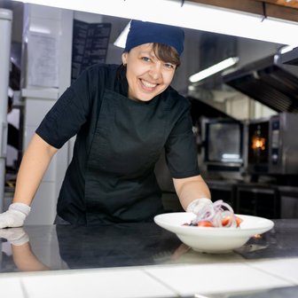 In einer professionellen Küche lächelt eine Köchin, während sie ein Gericht mit frischen Tomaten und Zwiebeln anrichtet.