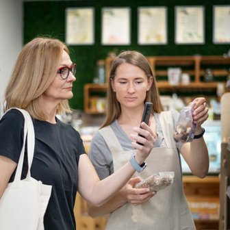 Zwei Frauen im Supermarkt vergleichen Snacks, eine von ihnen hat eine Stofftasche und die andere nutzt ihr Smartphone zur Recherche.