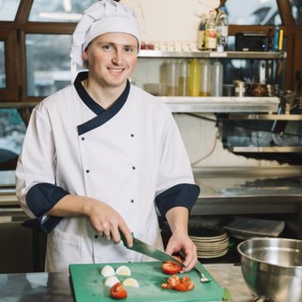 In einer gut ausgestatteten Küche bereitet ein lächelnder Koch Zutaten vor, während er Tomaten und Eier auf einem Brett schneidet.