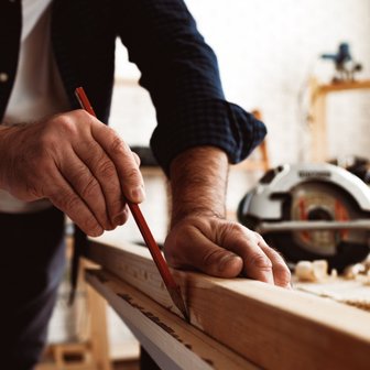 Ein Handwerker zeichnet präzise eine Linie auf ein Holzbrett, während eine Kreissäge im Hintergrund bereitsteht.