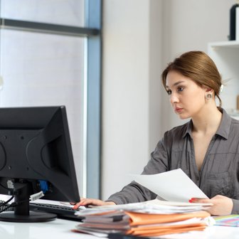 Am Schreibtisch einer hellen Büroumgebung studiert eine Frau aufmerksam Dokumente, während sie auf den Bildschirm tippt.