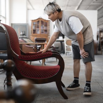 In einem hellen Raum mit antiken Möbeln untersucht ein Mann einen eleganten roten Sessel mit einem interessierten Blick.