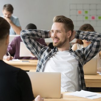 Ein lächelnder Mann mit Bart sitzt entspannt in einem Klassenzimmer, während andere konzentriert arbeiten.