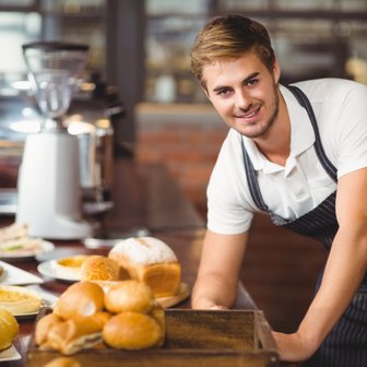 Ein junger Mann in einem gestreiften Schürze lächelt, während er frische Brötchen auf einem Holzbrett anrichtet.