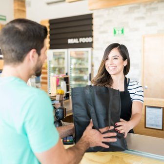 Eine lächelnde Kassiererin überreicht einem Kunden eine schwarze Tragetasche an der Kasse eines Lebensmittelgeschäfts.
