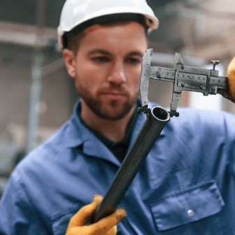 In einer Werkstatt prüft ein Arbeiter mit Handschuhen sorgfältig die Maße eines Rohres mit einem präzisen Messwerkzeug.