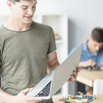 Ein junger Mann hält einen Laptop in der Hand und lächelt, während ein anderer Mann im Hintergrund an einem Tisch arbeitet.