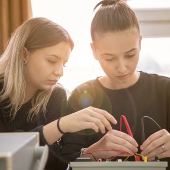 Die beiden Frauen zeigen Interesse und Teamarbeit, während sie an einem elektrischen Gerät mit verschiedenen Kabeln arbeiten.