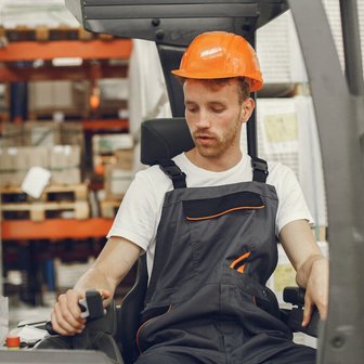 In einem Lagerhaus bedient ein Arbeiter in blauer Latzhose einen Gabelstapler, während er auf die Instrumente schaut.