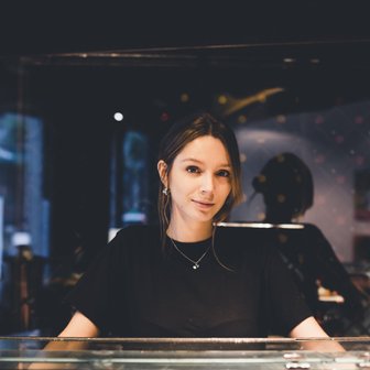 Eine junge Frau mit langen Haaren lächelt hinter einer Glasvitrine, die mit köstlichen Häppchen dekoriert ist.