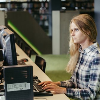 In einer Bibliothek arbeitet eine Frau an einem Computer, während im Hintergrund Regale mit Büchern sichtbar sind.